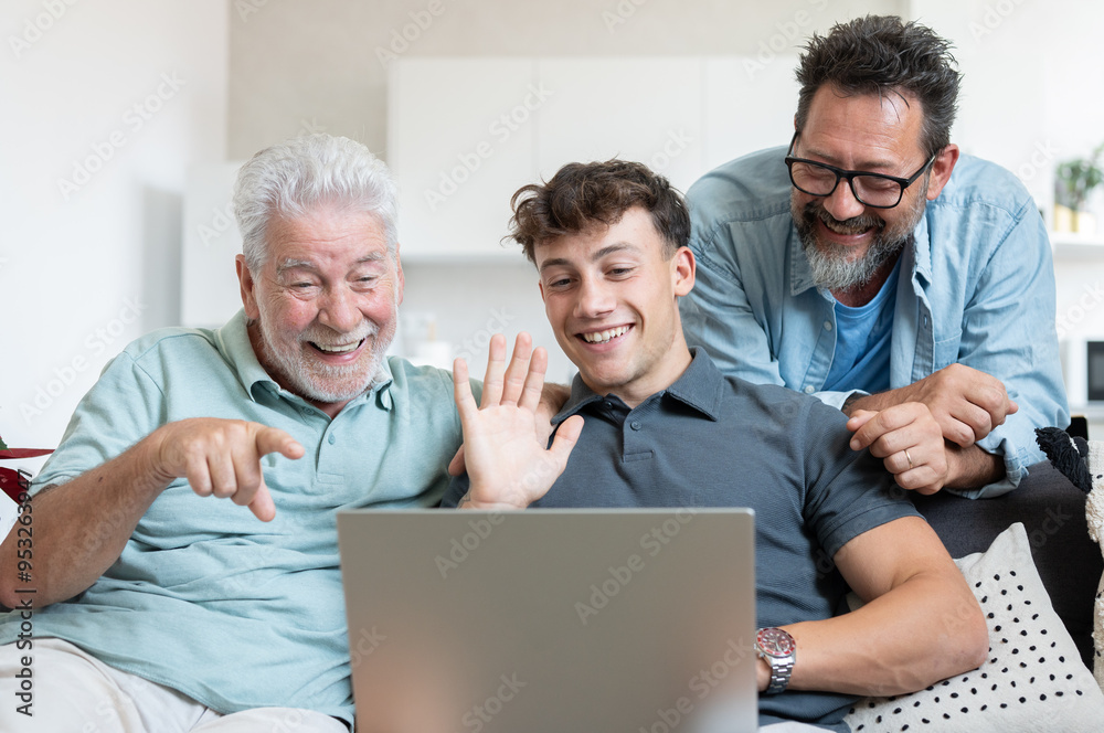 Happy multigenerational family of three males sit together on the couch smiling video calling by laptop. Grandfather, mature son and young grandson share positive moments in total relaxation Happy multigenerational family of three males sit together on the couch smiling video calling by laptop. Grandfather, mature son and young grandson share positive moments in total relaxation