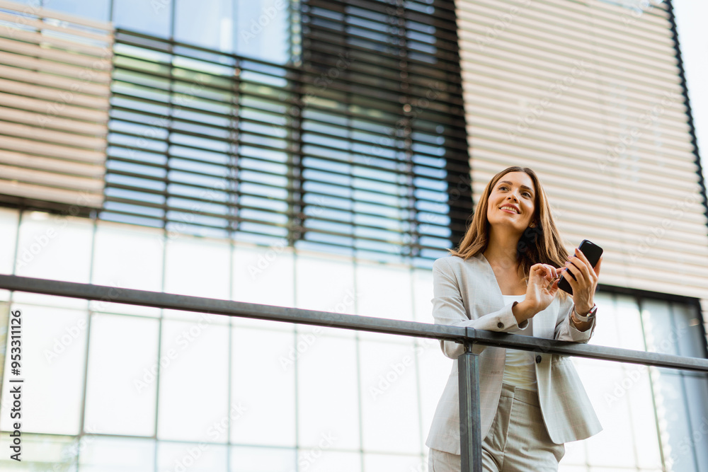 Fototapeta premium Smiling business woman enjoying a sunny day outdoors while texting on her smartphone near modern architecture in an urban setting