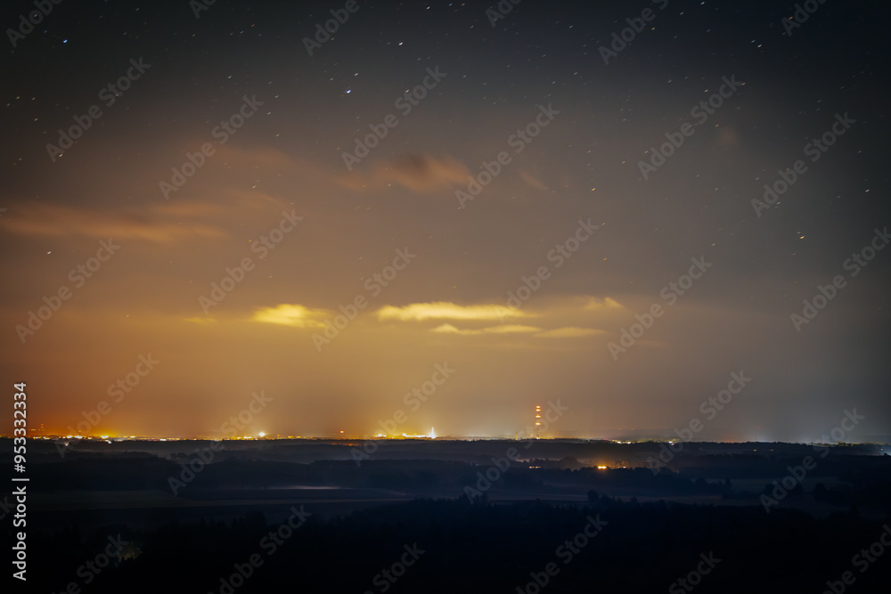 Obraz premium A serene night sky filled with stars stretches over a distant horizon illuminated by the soft glow of city lights. The contrast between the dark landscape and the warm light creates a peaceful