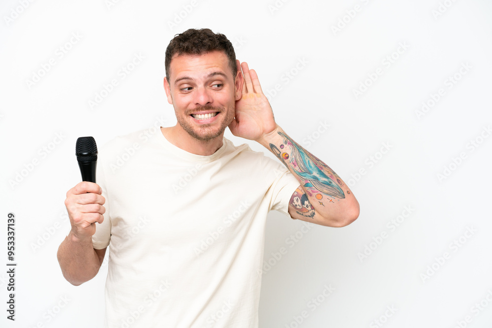 Young caucasian singer man picking up a microphone isolated on white background listening to something by putting hand on the ear Young caucasian singer man picking up a microphone isolated on white background listening to something by putting hand on the ear