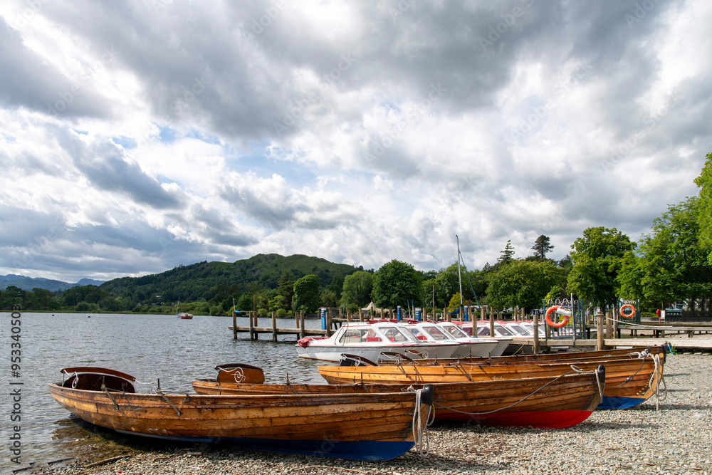 Small rowing boats and motor boats on pebbled beach of Lake Windemere in Lake District National Park near Ambleside, UK with a dramatic clouded and sunny sky Small rowing boats and motor boats on pebbled beach of Lake Windemere in Lake District National Park near Ambleside, UK with a dramatic clouded and sunny sky
