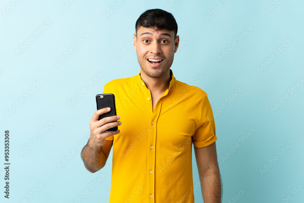 Young Colombian man using mobile phone isolated om blue background with surprise and shocked facial expression Young Colombian man using mobile phone isolated om blue background with surprise and shocked facial expression