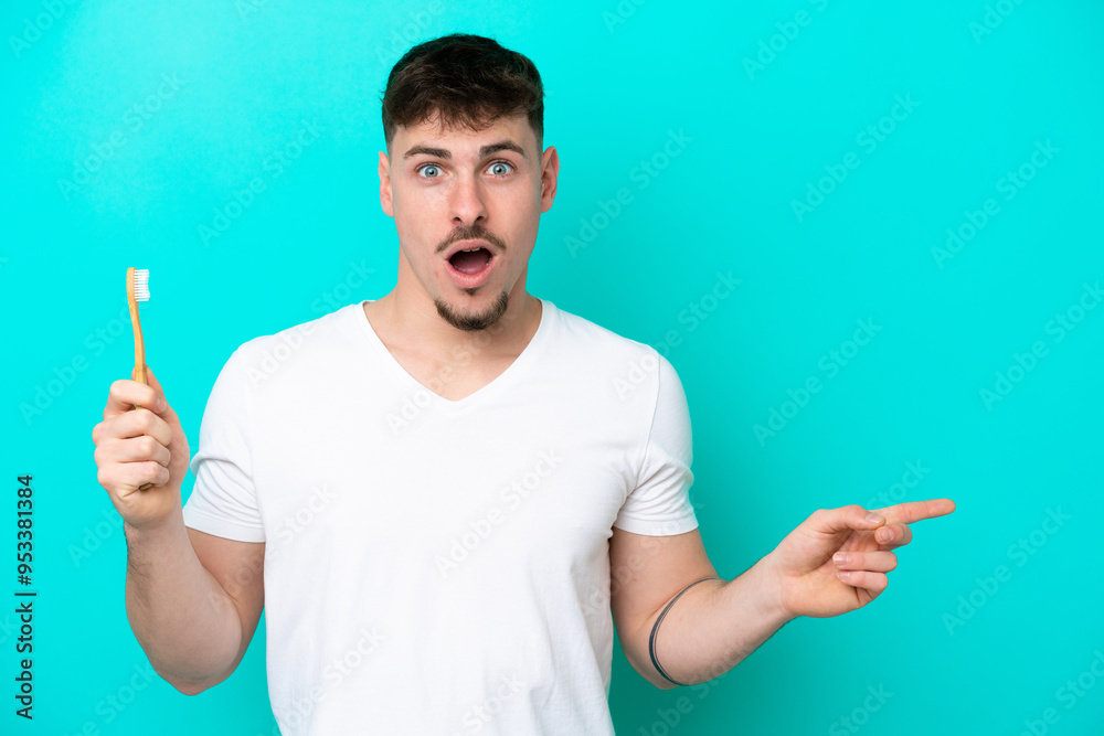 Young caucasian man brushing teeth isolated on blue background surprised and pointing side