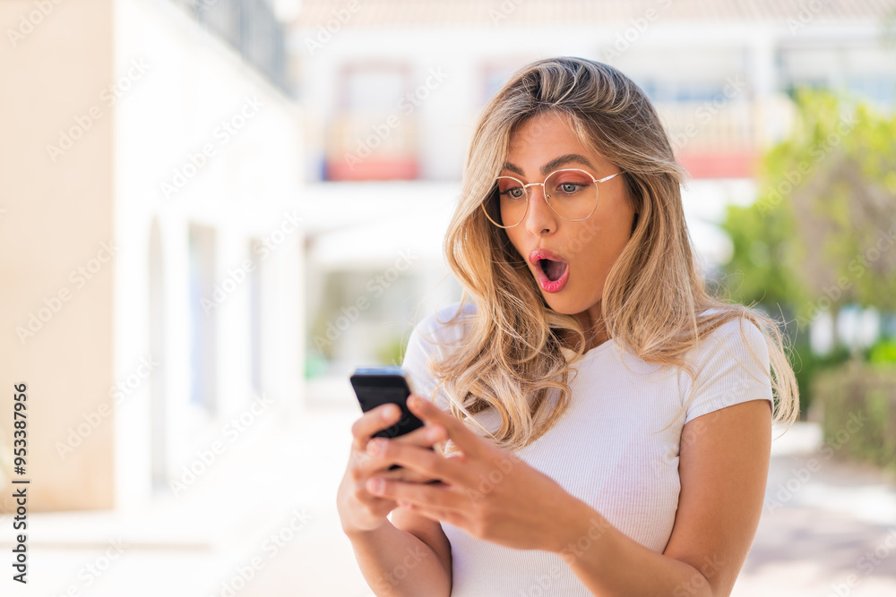 Pretty blonde Uruguayan woman looking at the camera while using the mobile with surprised expression Pretty blonde Uruguayan woman looking at the camera while using the mobile with surprised expression