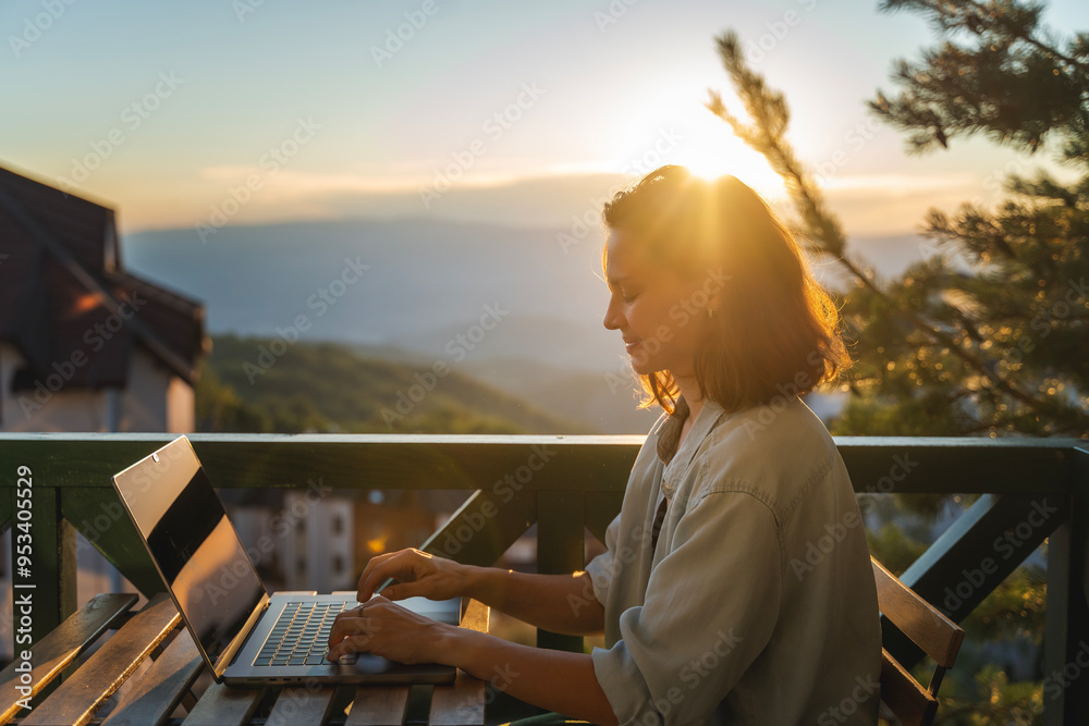 Young woman sitting on balcony with view of mountains and sunset working using laptop