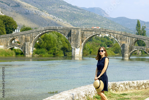 Attractive smiling woman in dress with hat in hands on summer day stands against the background of famous landmark of Trebinje - Arslanagić (Perovic) bridge. Female tourist in Bosnia and Herzegovina. photo