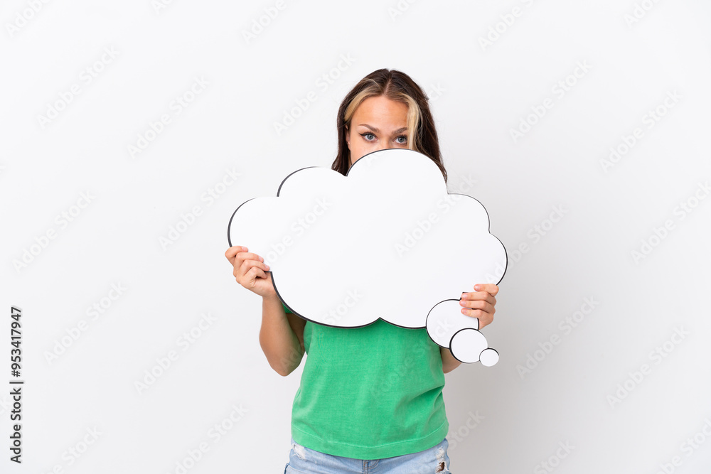 Teenager Russian girl isolated on blue background holding a thinking speech bubble and hiding behind it Teenager Russian girl isolated on blue background holding a thinking speech bubble and hiding behind it