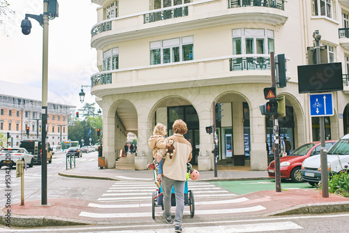Young father crossing busy road at crosswalk, holding toddler kid in his arms and pushing stroller, back view image