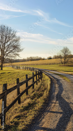 A winding, gravel road leads through a serene countryside, framed by a rustic wooden fence and golden fields bathed in the warm glow of the afternoon sun.