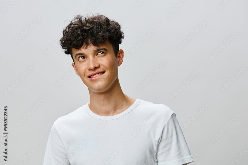 Portrait of a young man with curly hair wearing a white t shirt against a soft gray background, displaying a playful expression and thoughtful glance Portrait of a young man with curly hair wearing a white t shirt against a soft gray background, displaying a playful expression and thoughtful glance