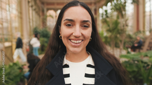 Confident young woman in navy coat and striped shirt standing in lush greenhouse photo