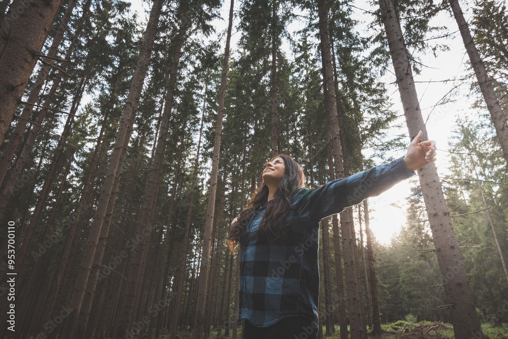 © Fauren - Happy woman in a plaid shirt with outstretched arms enjoying the serenity of a dense forest, surrounded by tall trees, feeling freedom and connection with nature
