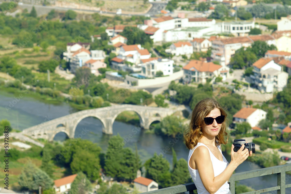 An attractive young woman stands with a DSLR camera in her hands, about to take a photo from the observation deck of the famous Trebinje landmark - the Arslanagic Bridge (Bosnia and Herzegovina). An attractive young woman stands with a DSLR camera in her hands, about to take a photo from the observation deck of the famous Trebinje landmark - the Arslanagic Bridge (Bosnia and Herzegovina).
