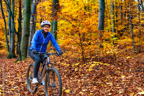 Middle aged woman wearing blue sporty jacket, black cycling pants and white bike helmet riding bicycle in city forest in autumn scenery. Front view.