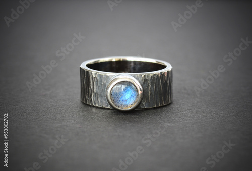 Hand made sterling silver ring with natural blue shine labradorite round gemstone in bezel setting. Darkened oxidised silver hammered ring with blue gemstone on gray paper gradient background.