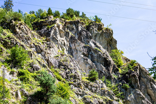Rocky cliffside with greenery and blue sky near a power line on a sunny day in a mountainous region