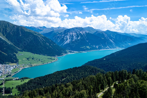 A panoramic view of turquoise lake surrounded by mountains and lush greenery on a sunny day in the Alps