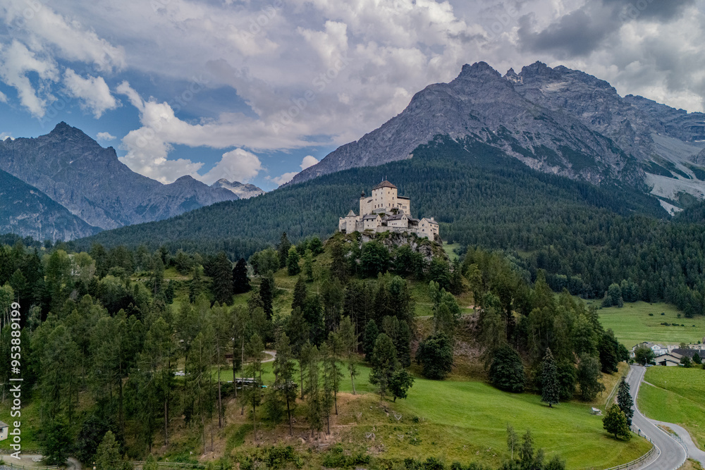 Aerial view of a castle perched on a hill surrounded by lush forests and towering mountains during a cloudy day Aerial view of a castle perched on a hill surrounded by lush forests and towering mountains during a cloudy day