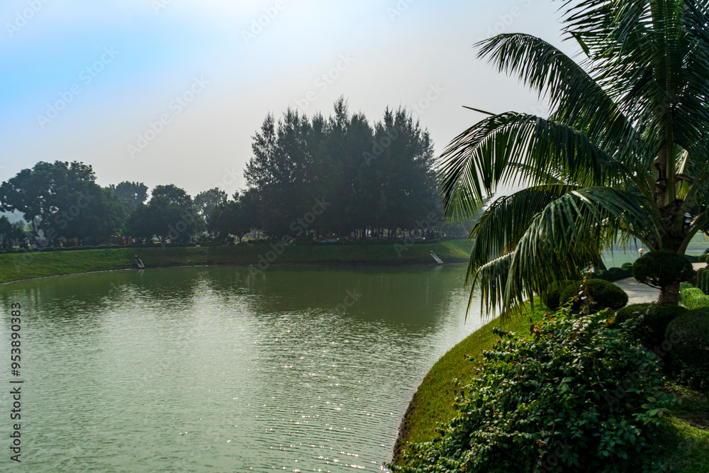 Obraz premium serene lake surrounded by lush greenery and palm trees under hazy sky.