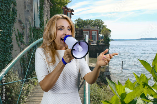 Happy cheerful blonde woman giving a speech in megaphone outdoors. Summer lake landscape. photo