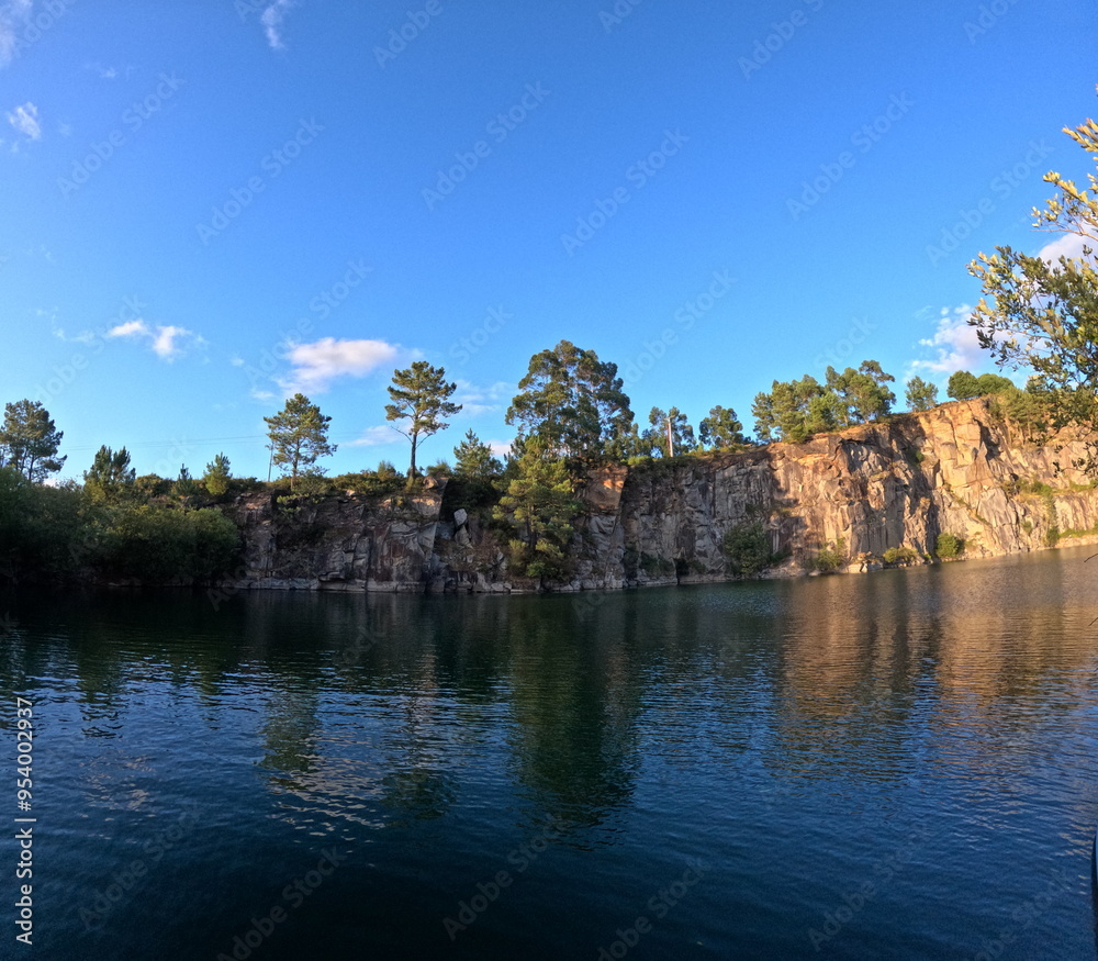 The image depicts the tranquil scene of the Catoira quarries, where steep rocky cliffs adorned with pine trees rise above a calm, reflective body of water. The sky is clear and blue, enhancing the ser The image depicts the tranquil scene of the Catoira quarries, where steep rocky cliffs adorned with pine trees rise above a calm, reflective body of water. The sky is clear and blue, enhancing the ser