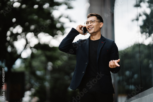 Confident Businessman on the Phone: A young man in a stylish black blazer and glasses walks confidently in the city, engrossed in a phone conversation. His smile suggests a successful deal or exciting