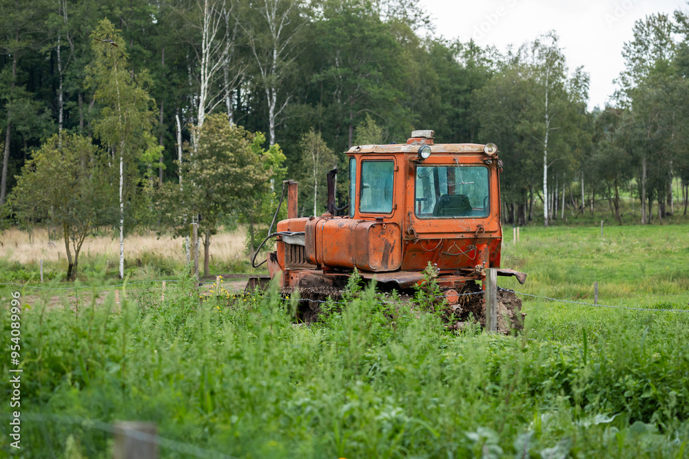 Fototapeta premium An old orange tractor parked in a field overgrown with tall grass, with trees in the background.