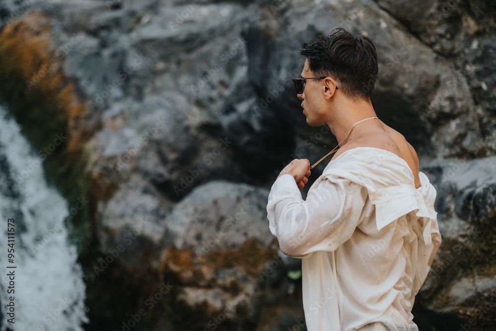 Young man standing by a rocky waterfall. Wearing sunglasses and a wet white shirt, he appreciates the beauty of nature and adventure. Young man standing by a rocky waterfall. Wearing sunglasses and a wet white shirt, he appreciates the beauty of nature and adventure.