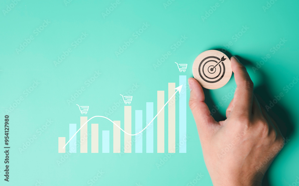 A hand holding a wooden with a target icon, goal achievement or strategic planning, chart and shopping cart icons business growth, an upward trending line indicating positive financial performance.