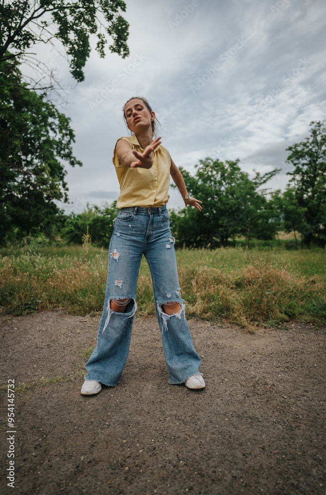 A young woman dressed in ripped jeans and a yellow shirt spends time outdoors, appreciating the serene natural environment under a cloudy sky. A young woman dressed in ripped jeans and a yellow shirt spends time outdoors, appreciating the serene natural environment under a cloudy sky.