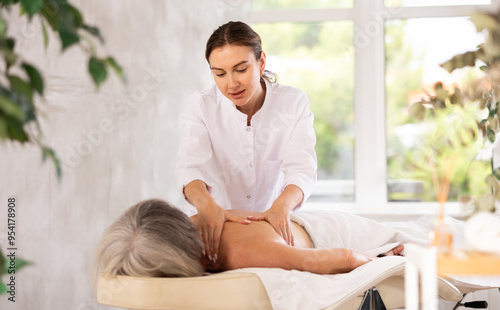 Quadro su tela Young woman masseur performs shoulder stroking and patting during massage session for senior woman client