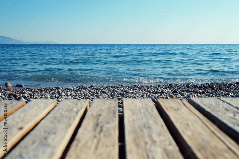 Fototapeta premium A tranquil seascape featuring a calm blue ocean meeting a pebbled shore, viewed from a wooden pier under a clear sky.