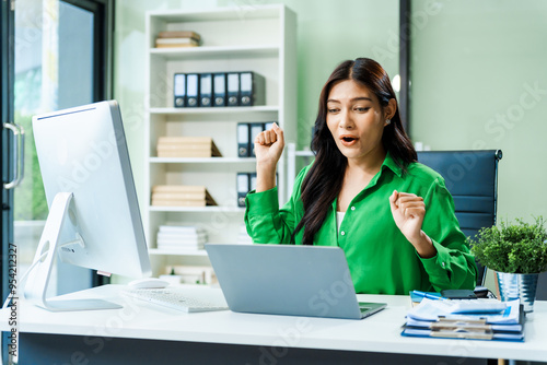 successful and happy young Asian businesswoman in a formal suit works at her desk in a modern glass office. consults on business strategies LGBTQ inclusivity, managing multiple projects efficiently.