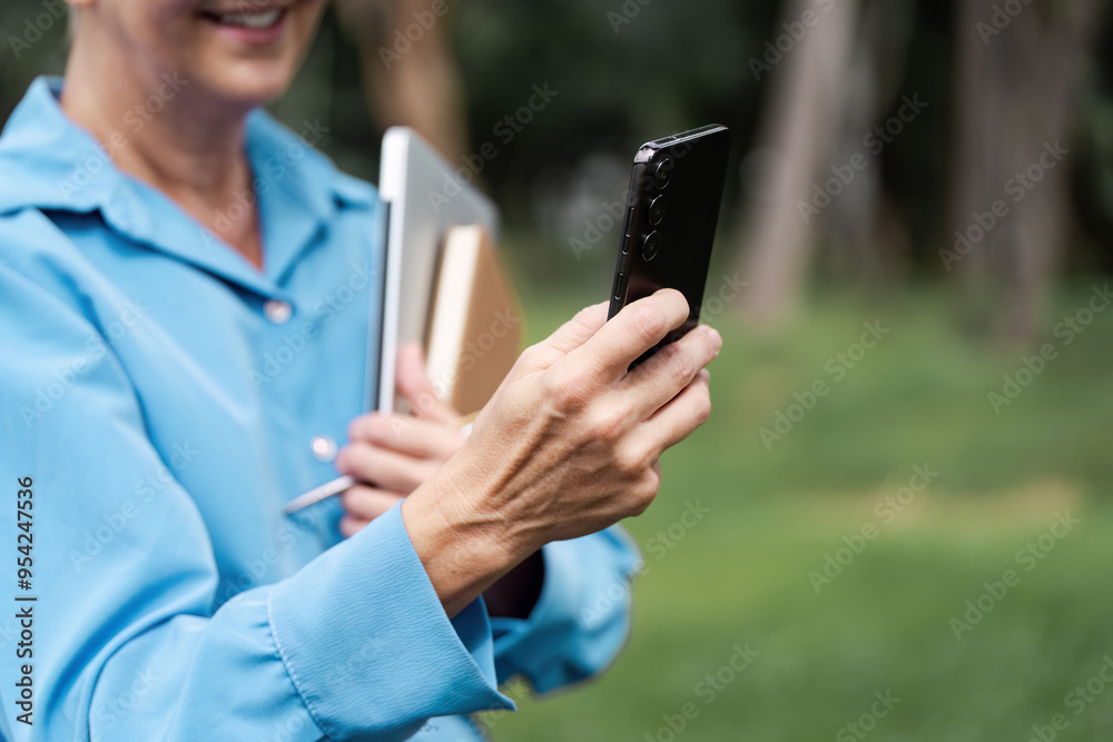 Senior Woman Using Smartphone Outdoors While Holding Books and Tablet in a Park Setting Senior Woman Using Smartphone Outdoors While Holding Books and Tablet in a Park Setting