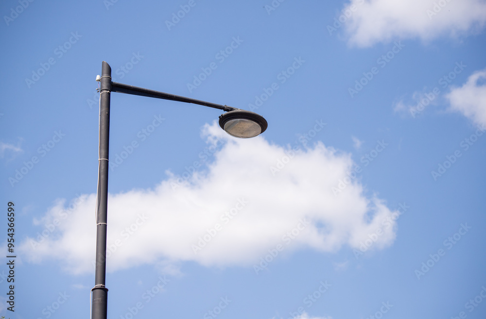 A solitary streetlamp stands against a bright blue sky filled with fluffy white clouds during the daytime A solitary streetlamp stands against a bright blue sky filled with fluffy white clouds during the daytime