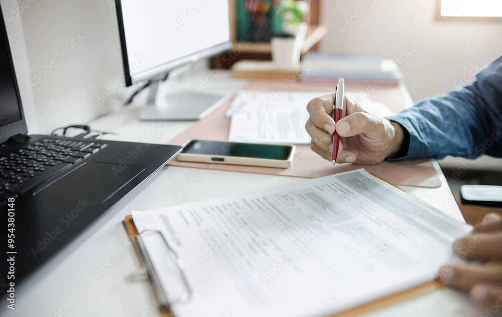 Asian businessman working on desk with laptop and desk top PC holding pen checking business contract and document report before sign Asian businessman working on desk with laptop and desk top PC holding pen checking business contract and document report before sign