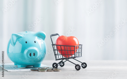 A blue piggy bank, a pile of coins, and a red heart in a shopping cart sit on a table against a bright white background. The image symbolizes saving money for health care or financial well-being. photo