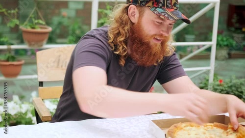 A red-bearded curly-haired guy opens a pizza box and takes out a fresh piece with cheese, biting off with pleasure, satisfying his hunger at a table outside. photo