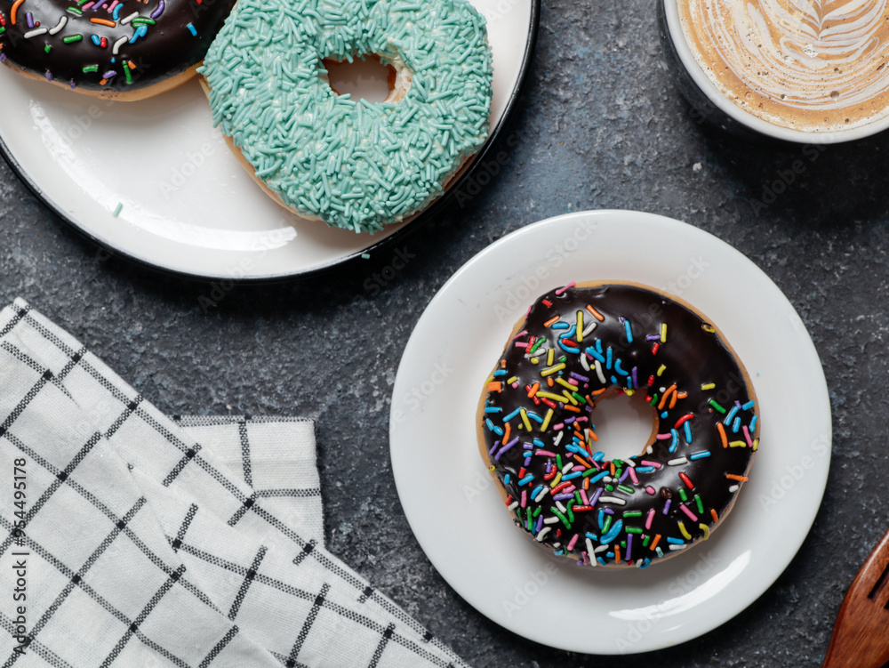 An assortment of donuts is arranged on two plates on a dark, textured surface. A chocolate donut with colorful sprinkles is on a white plate, while another plate has donuts with green sprinkles An assortment of donuts is arranged on two plates on a dark, textured surface. A chocolate donut with colorful sprinkles is on a white plate, while another plate has donuts with green sprinkles