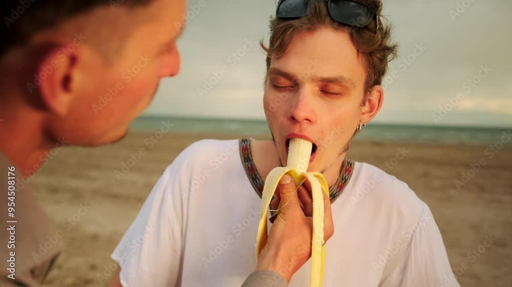 Slow motion, two cute guys eating banana romantically looking into each other's eyes on the beach. Close up. Slow motion, two cute guys eating banana romantically looking into each other's eyes on the beach. Close up.