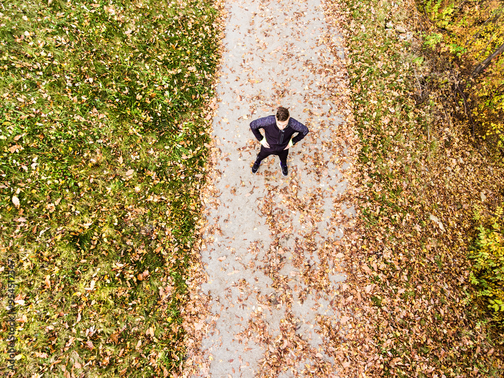 Fototapeta premium Aerial view of a runner standing on jogging path with fallen leaves and resting after in autumn park. Morning running training.