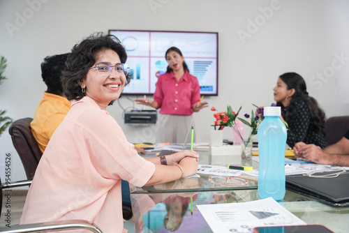 Happy young indian woman sitting around a table with colleagues smiling at the camera during meeting. Woman giving presentation in office using tv screen charts, graphs and statistics, people and work photo