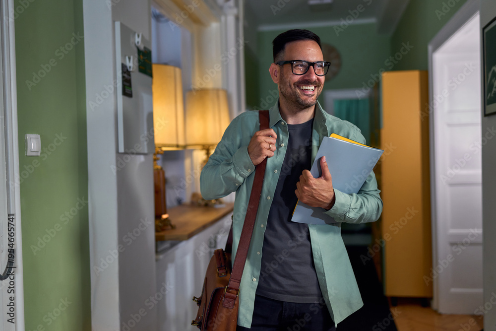 Fototapeta premium A man with glasses, wearing a green shirt and carrying a shoulder bag and folders, smiles while standing in a well-lit hallway.