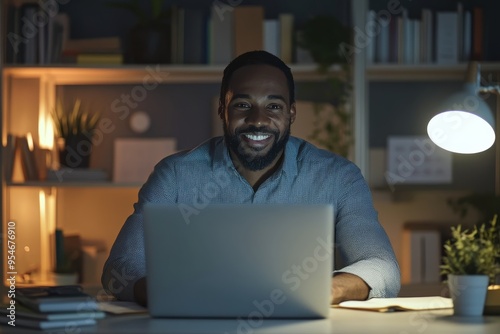 A happy Black man is sitting at his desk in front of an open laptop, working on a digital marketing project late at night, with bookshelves behind him and soft lighting illuminating the scene.