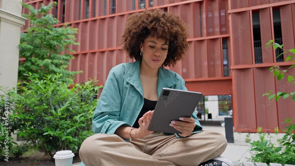 Camera focus on African-American girl sitting on bench in middle of small green zone for relaxation. Using tablet device. Scrolling through everything on screen. Searching for product or information.
