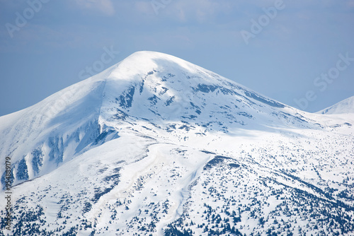 Snow-covered mountain peak under clear blue sky, with patches of snow on slopes and scattered trees. Serene winter landscape contrasts sharply with the dark, forested areas below. Hoverla, Ukraine photo