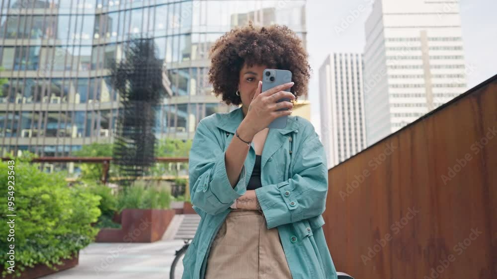 Curly girl using modern smartphone while spending time outside. Watching funny video on phone. Cheerfully smiling. Standing in middle of street. Skyscrapers located behind happy female. Curly girl using modern smartphone while spending time outside. Watching funny video on phone. Cheerfully smiling. Standing in middle of street. Skyscrapers located behind happy female.