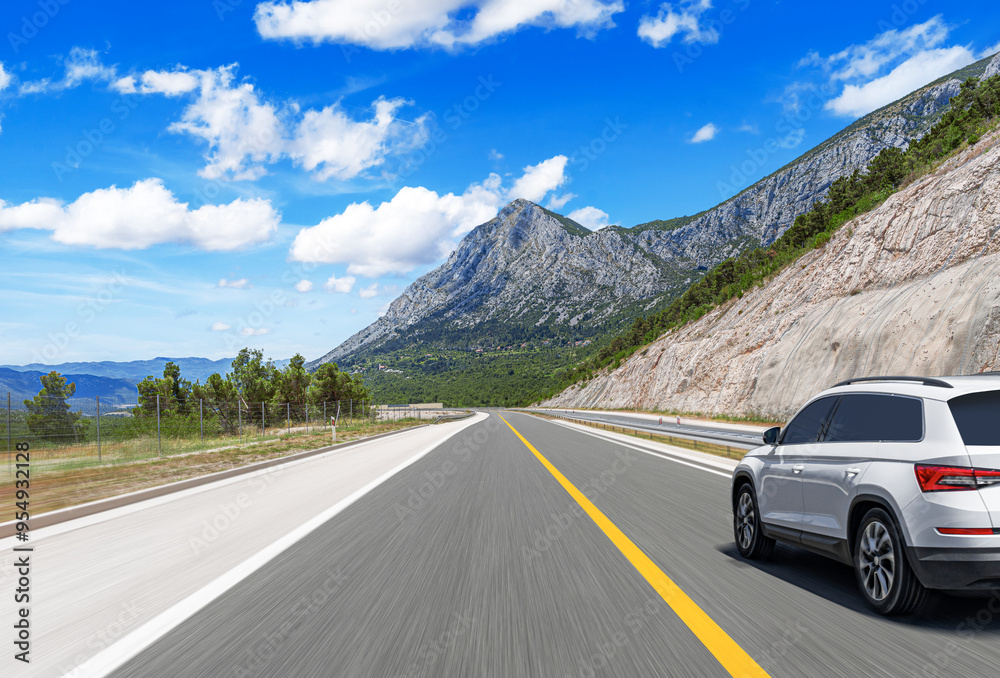 A white car drives along the highway against the backdrop of rocky mountains on a sunny day.