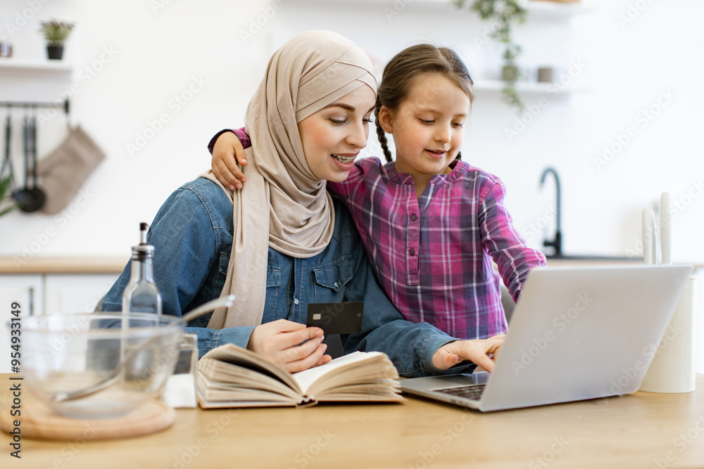 Happy family cooking together, embracing technology for home activities. Muslim mother and daughter sitting at kitchen table and buying ingredients for meal using laptop, bank card and recipe book. Happy family cooking together, embracing technology for home activities. Muslim mother and daughter sitting at kitchen table and buying ingredients for meal using laptop, bank card and recipe book.