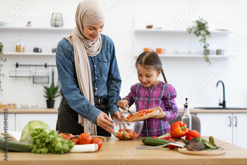 Happy family bonding while cooking fresh and healthy meal in modern kitchen. Parent and child teamwork. Muslim mother and daughter pour chopped vegetables from wooden board into salad bowl.